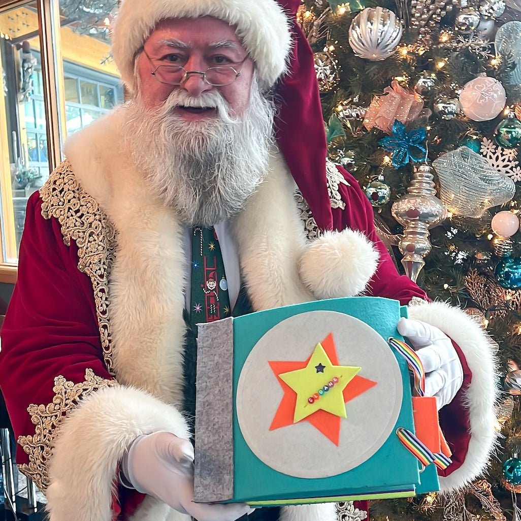 Person dressed as Santa Claus holding a quiet book in front of a decorated Christmas tree.